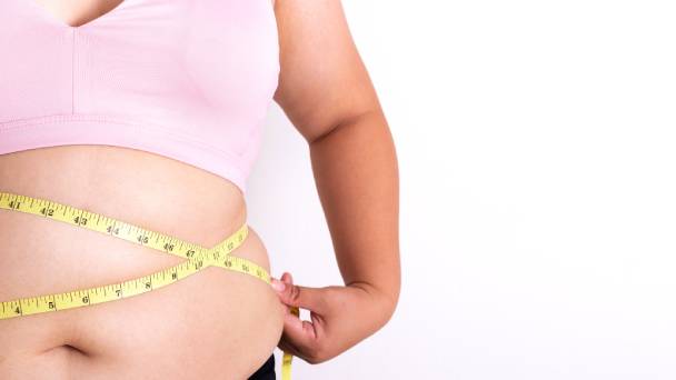 Corpo de uma mulher obesa. Ela vese um top rosa e segura uma fita métrica em torno da cintura